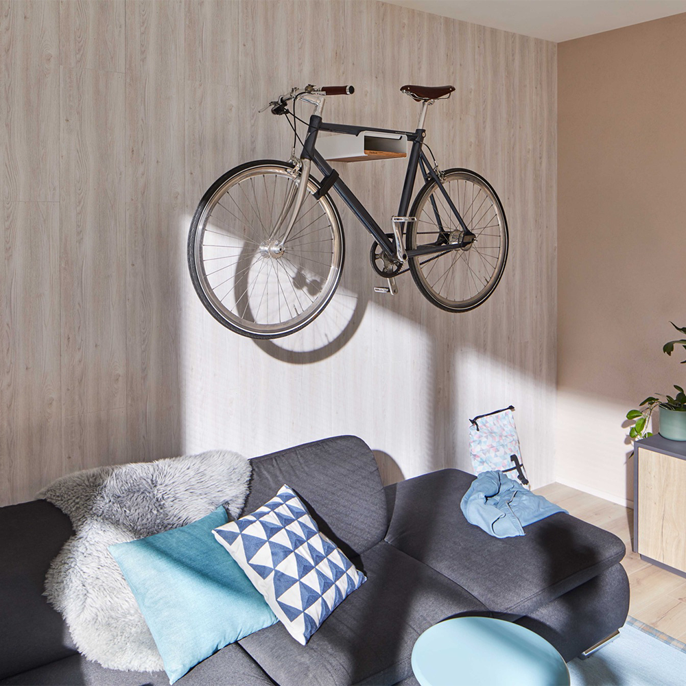 Blaues Fahrrad liegt auf weißer Fahrrad Wandhalterung mit Walnuss Einlegebrett vor grauer Wand und über grauem Sofa mit Kissen